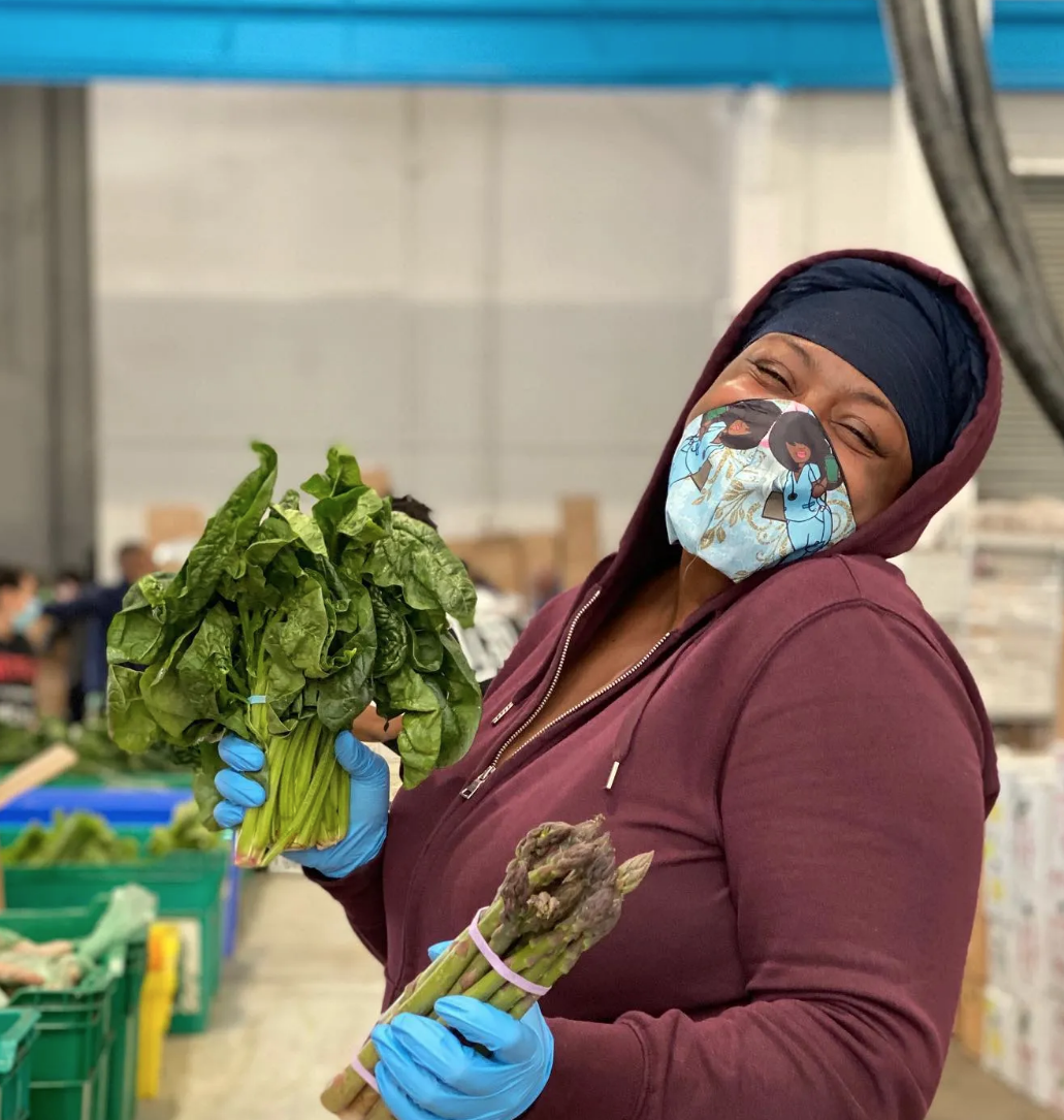 Woman holding produce in a warehouse. She is smiling behind her face mask.